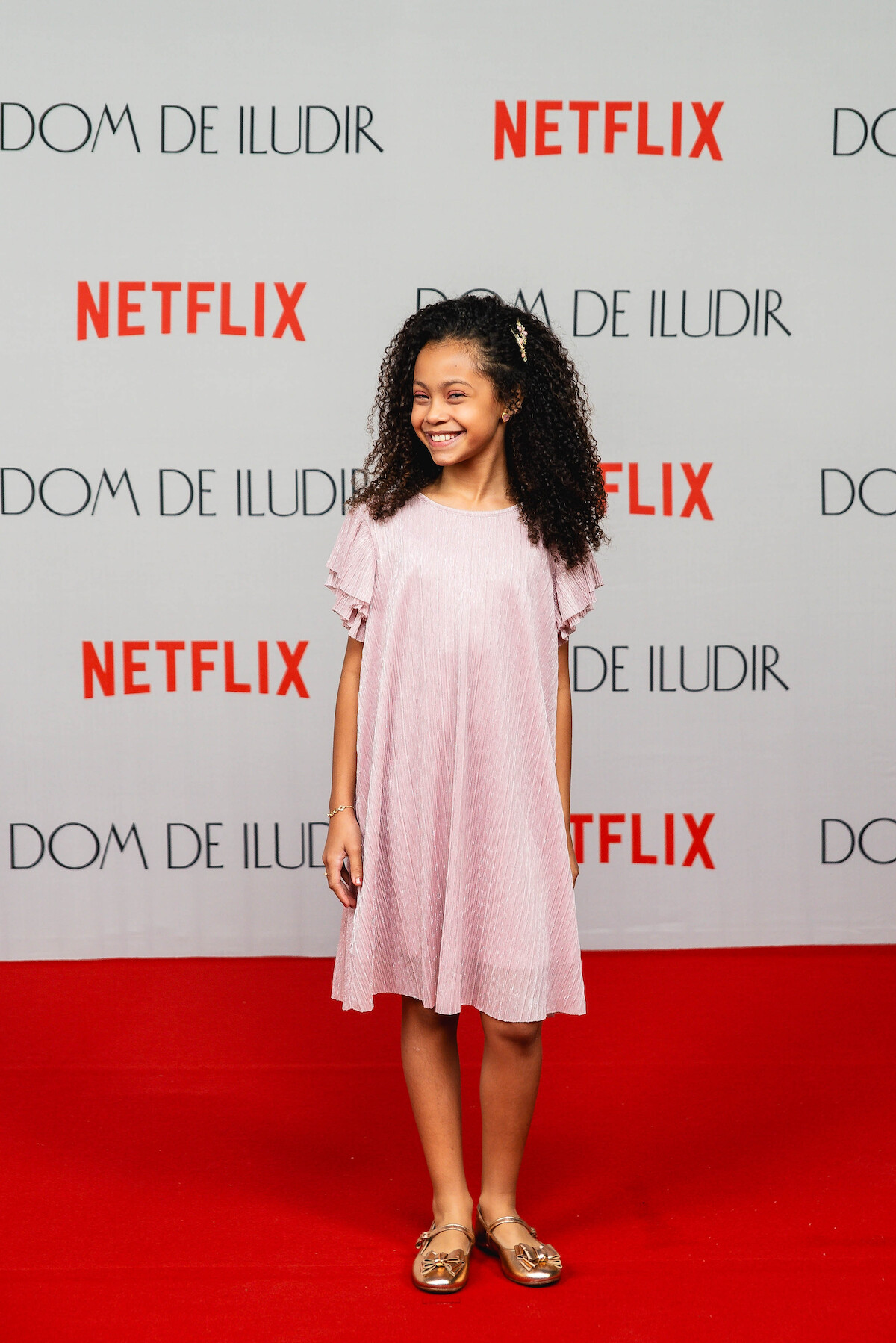 A atriz Laura Vick é uma menina negra de cabelos encaracolados. Ela veste um vestido rosa e sapatos dourados