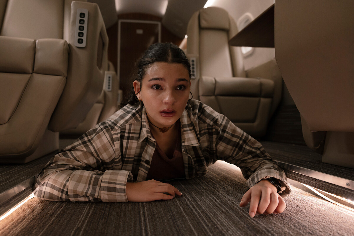 Young woman in plaid shirt crawls on the carpeted floor of a private jet, looking distressed. The interior has beige leather seats and soft lighting, suggesting an urgent or dramatic situation inside an upscale aircraft cabin.