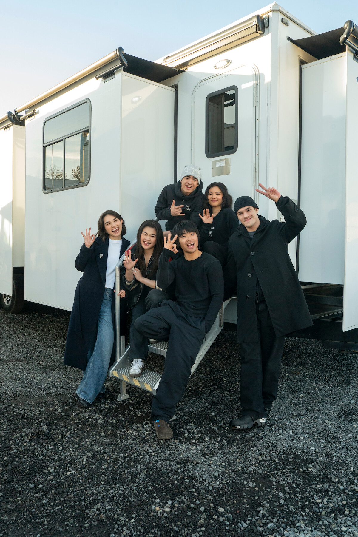 Six people posing and smiling in front of a white trailer on a gravel lot, wearing casual and dark jackets and coats, with a clear sky in the background, suggesting a friendly, outdoor, behind-the-scenes or film set environment.