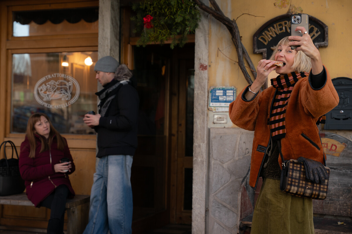 Woman taking a selfie and eating, smiling in front of a rustic trattoria. Two people, one seated with coffee, one standing, dressed warmly outside the restaurant. Relaxed, social outdoor cafe atmosphere.