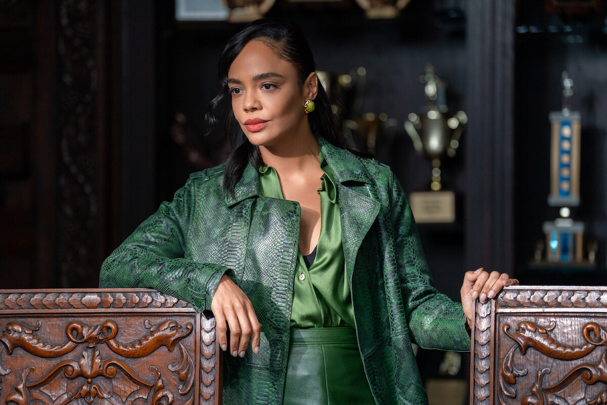 Woman in a green snakeskin jacket stands in an ornate room with carved wood chairs and trophy shelves.