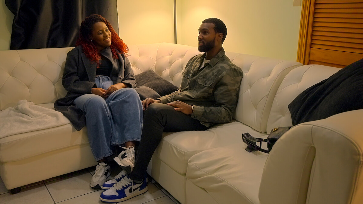 A woman and man sit talking on a cream-colored sectional sofa in a cozy living room with soft lighting, casual clothes, and sneakers, surrounded by pillows and blankets.