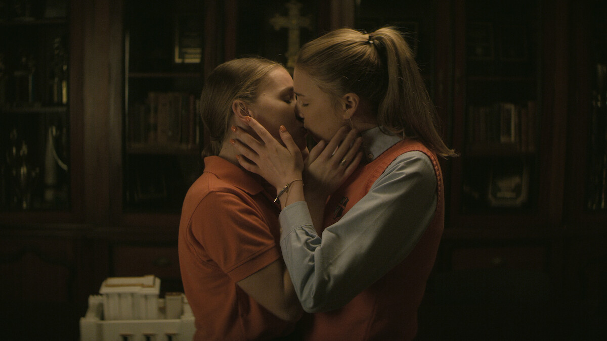 Two women in uniforms share a kiss in a dimly lit room with wooden bookshelves, religious artifacts, and a cross in the background, suggesting a school or religious setting.