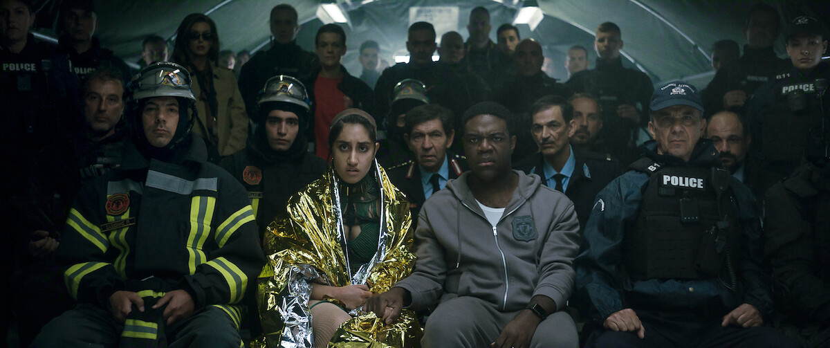 A diverse group of people, including police officers, firefighters, and civilians, sit closely together in what appears to be a dimly lit emergency shelter or tunnel, facing forward with serious and contemplative expressions.