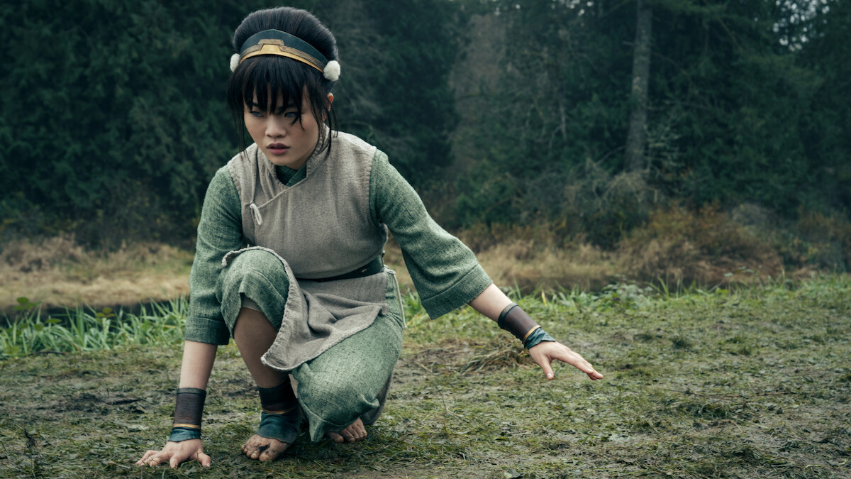 Miyako as Toph in traditional green and beige clothing crouches on grassy ground, with a focused expression, surrounded by a forest with tall trees and overcast lighting.