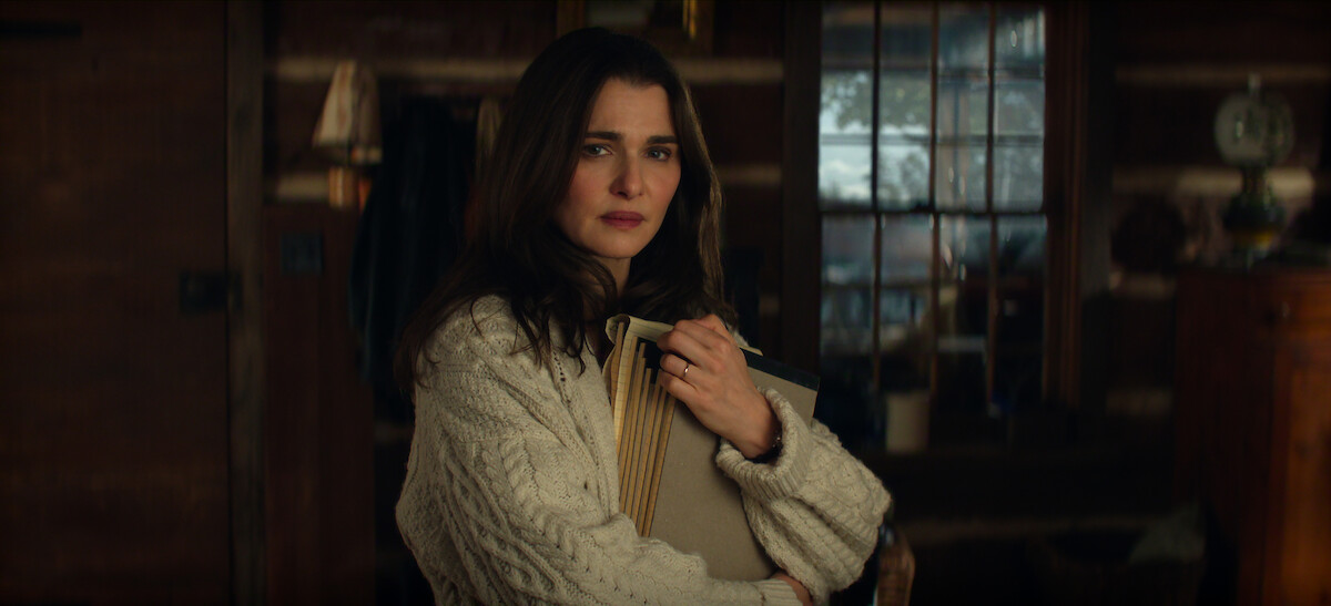 Woman in a cozy sweater holding books, standing indoors in a warmly lit rustic cabin with wooden walls and a window showing a snowy outdoor scene.