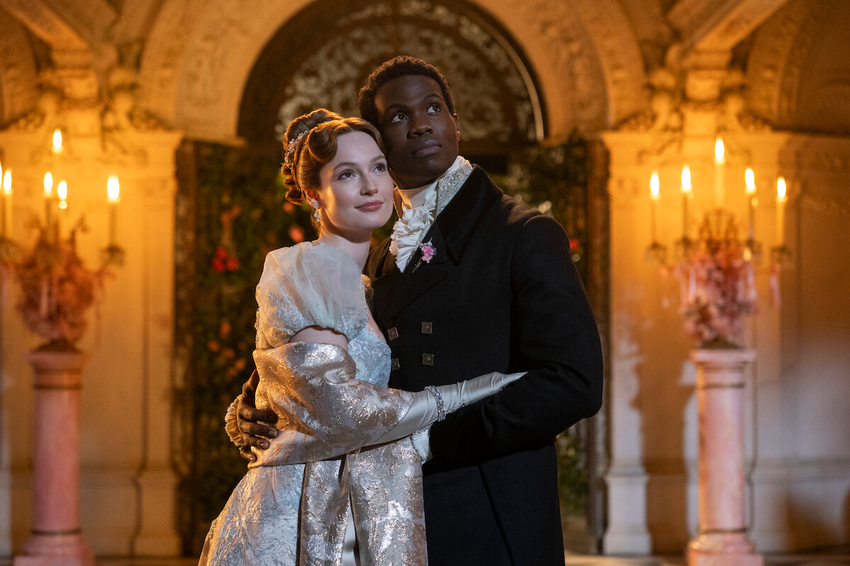 A couple in elegant historical attire embrace in a grand, ornately decorated ballroom with chandeliers and candlelight, creating a romantic and formal atmosphere.