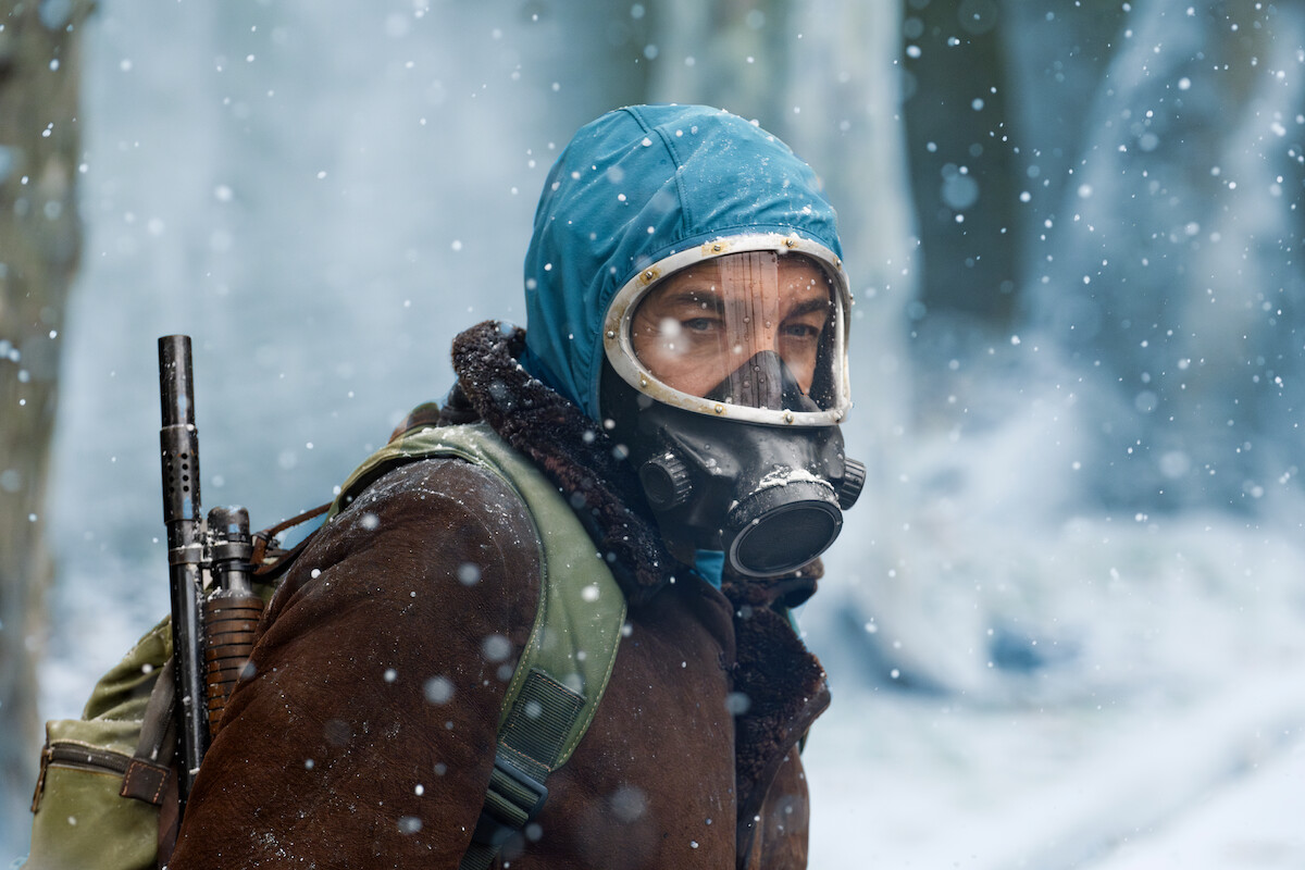 A person wearing a gas mask, a blue hood, and a brown jacket, standing in a snowy environment with snowflakes falling and a rifle strapped to their back.