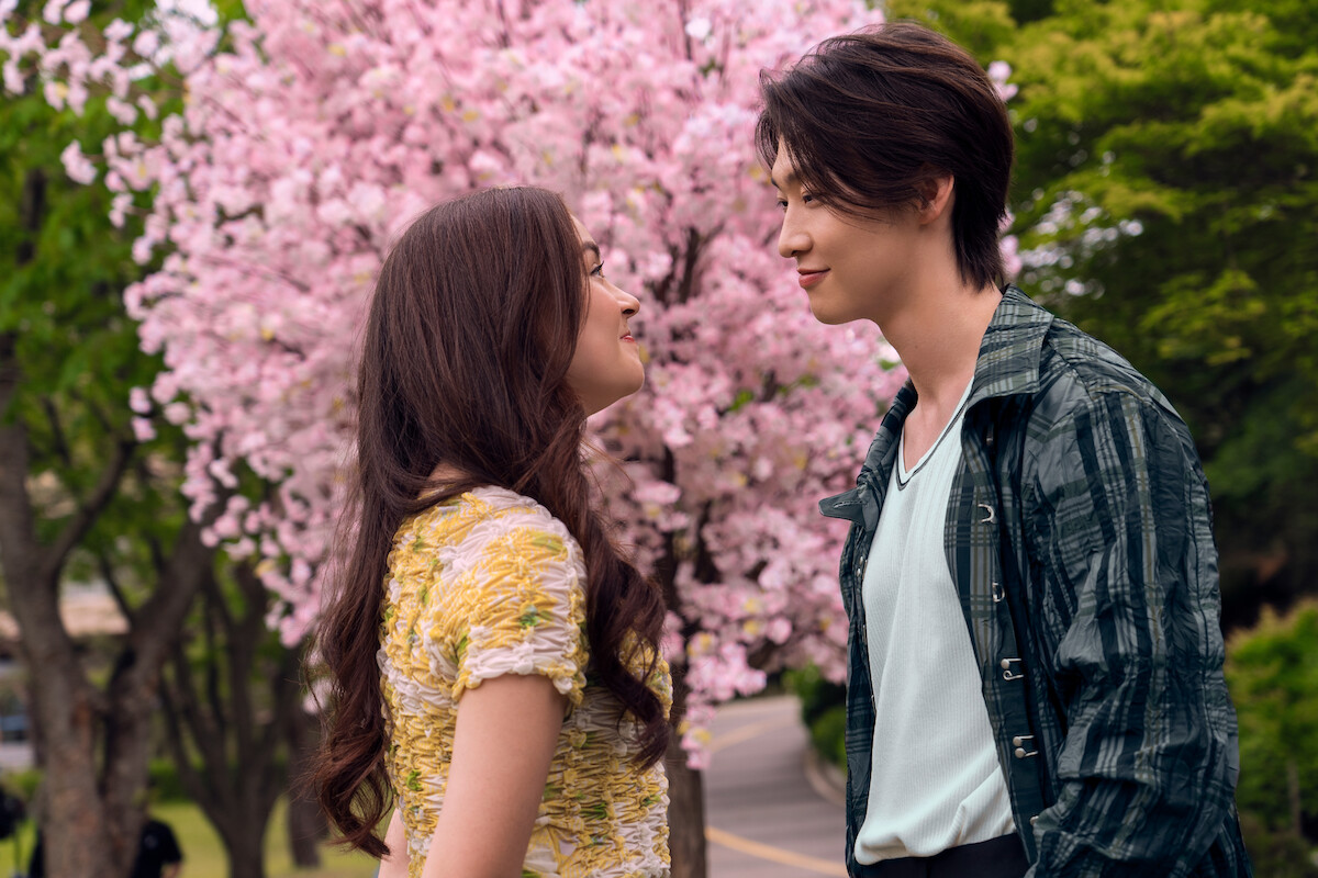 A young couple faces each other, smiling, in a park with blooming pink cherry blossom trees and green foliage in the background on a sunny day.