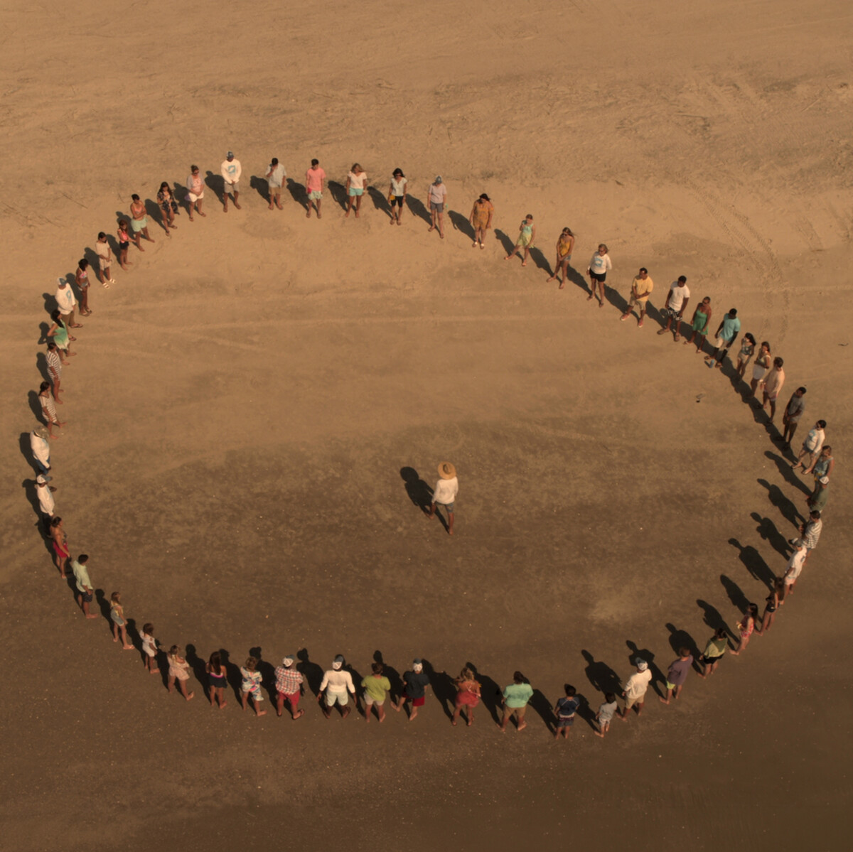 A group of people standing on sandy ground form a large circle around one person in the center, seen from above in a wide, open outdoor environment.