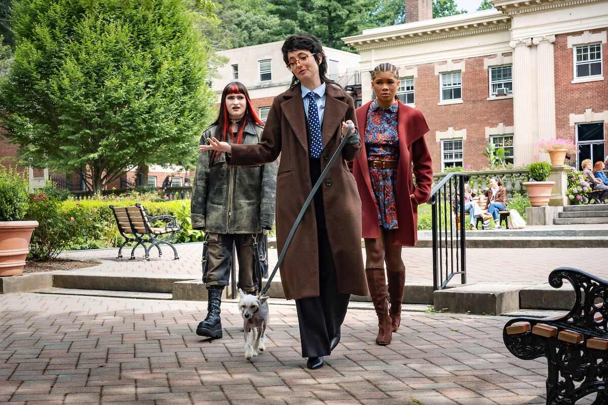 Three people walking outside on a brick path; one walks a small dog, in front of a red brick building with white columns, surrounded by greenery and benches, on a sunny day.