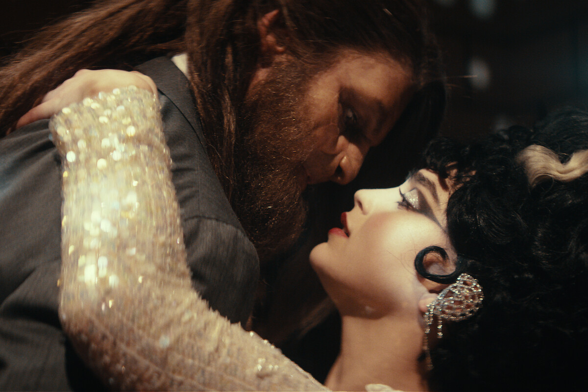 A man in a suit with a thick beard and a woman in glamorous attire with dramatic makeup and earrings share an intense, intimate dance in a dimly lit, elegant setting.