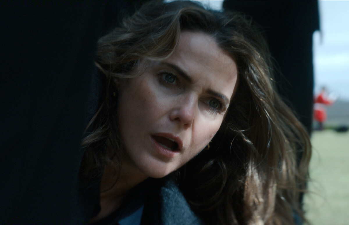 A woman with long hair looks concerned as she peers out from a dark, shadowy area outdoors, with blurred figures and a grassy field visible in the background.