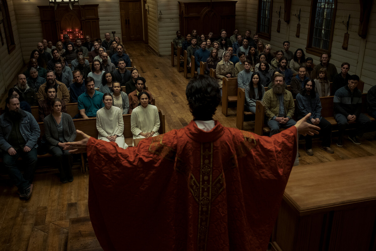 A priest in ornate red robes stands with arms open facing a full congregation in a wooden church, stained glass windows on the side, and attentive people seated on pews during a service.