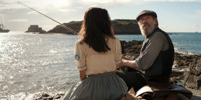 (L to R) Aria Mia Loberti as Marie-Laure, Hugh Laurie as Etienne LeBlanc. a woman sitting next to a fisherman