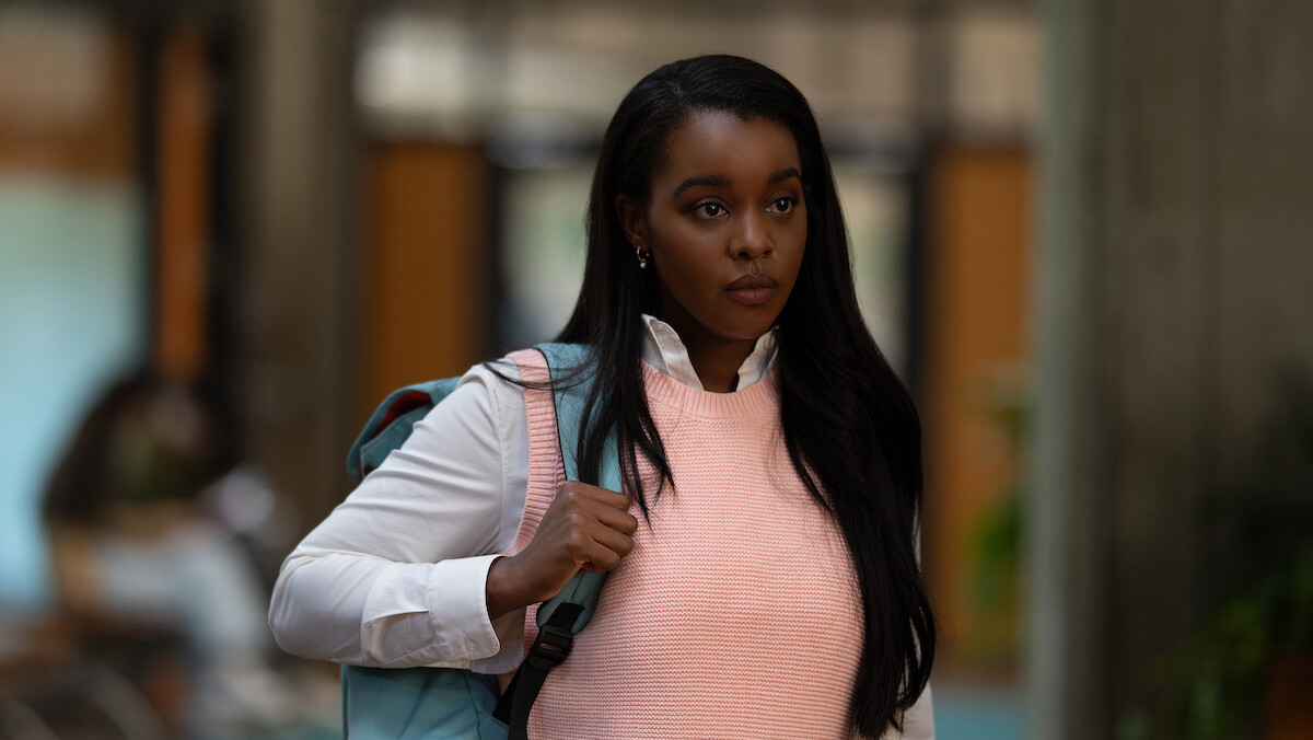 A young woman with long dark hair, wearing a pink vest and white shirt, stands indoors with a backpack strap over her shoulder, looking forward in a modern building with blurred background.