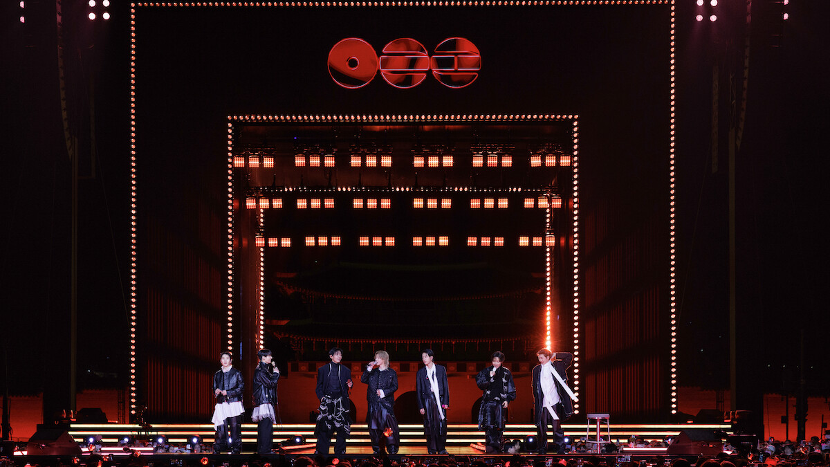SUGA, JIMIN, JIN, JUNG KOOK, V, J-HOPE, and RM on a brightly lit stage with orange and yellow lights, geometric decorations, and an audience watching a night concert.