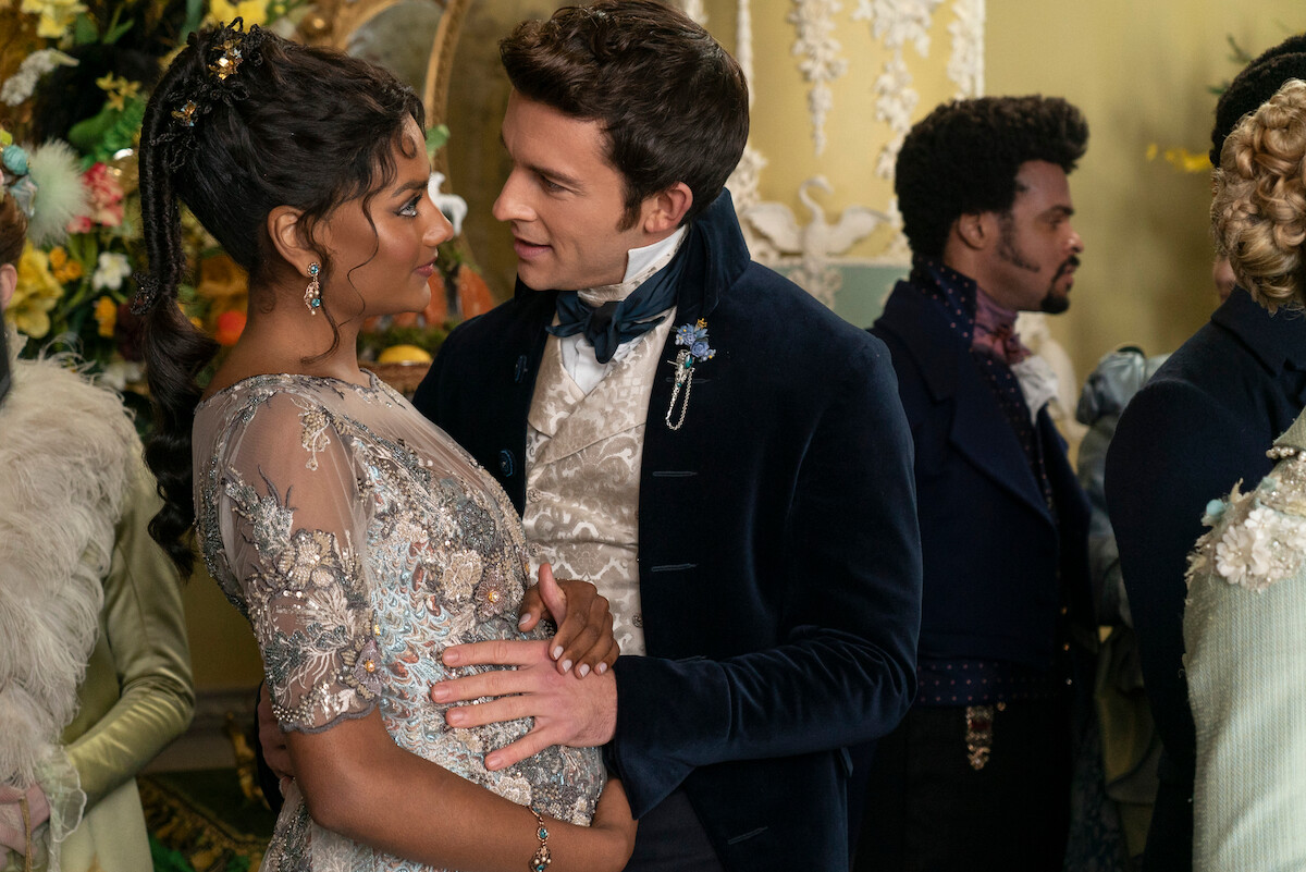 A couple in elegant period costumes share an intimate moment at a lavish indoor party, surrounded by well-dressed guests, floral decorations, and ornate furnishings.