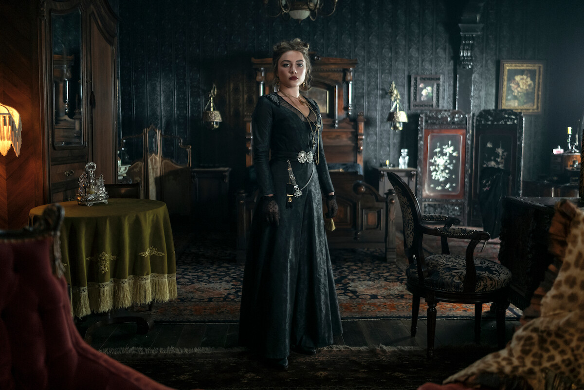 Woman in Victorian-era black dress stands in an ornate, dimly lit parlor filled with antique furniture, patterned rugs, and decorative objects, evoking a historical and dramatic atmosphere.