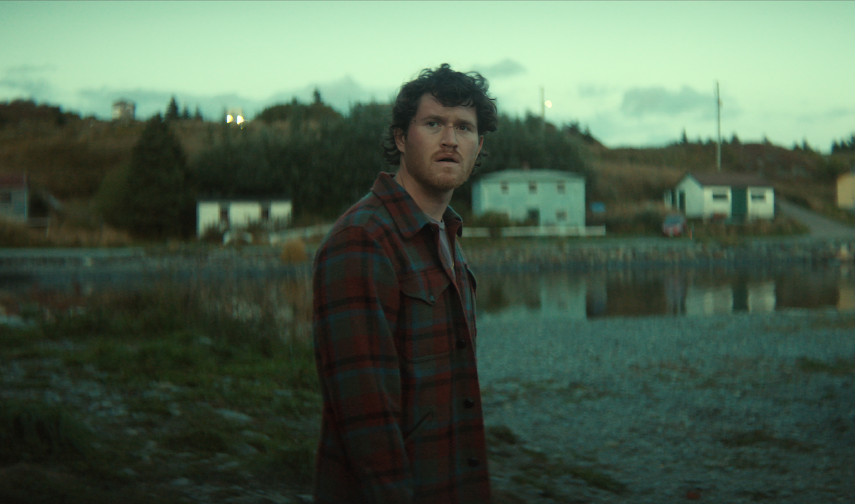 A man in a plaid jacket stands outdoors near a quiet shoreline, looking off into the distance with a concerned expression; small houses and trees are visible in the background under an overcast sky.