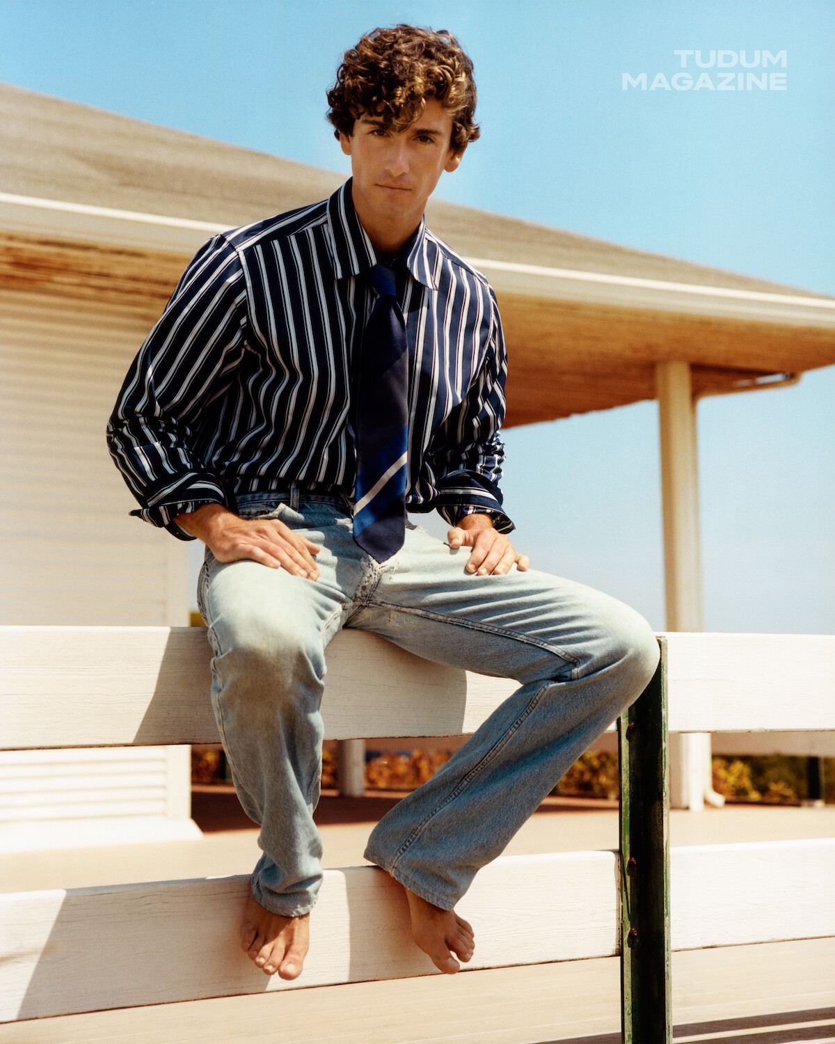A man in a striped shirt, tie, and jeans sits barefoot on a white fence outside a beach house on a sunny day, with "TUDUM MAGAZINE" text visible in the sky.