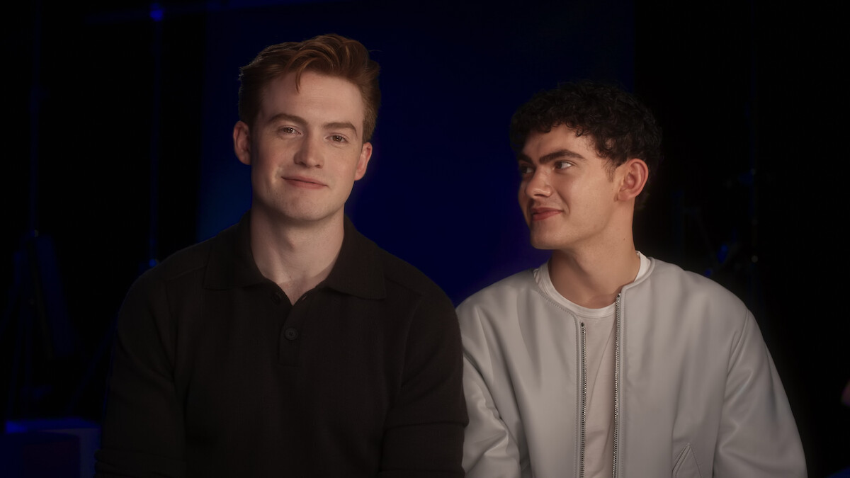 Two young men sitting closely together in a dimly lit indoor setting, one in a dark shirt smiling calmly at the camera while the other, in a light jacket, looks at him with a fond expression.