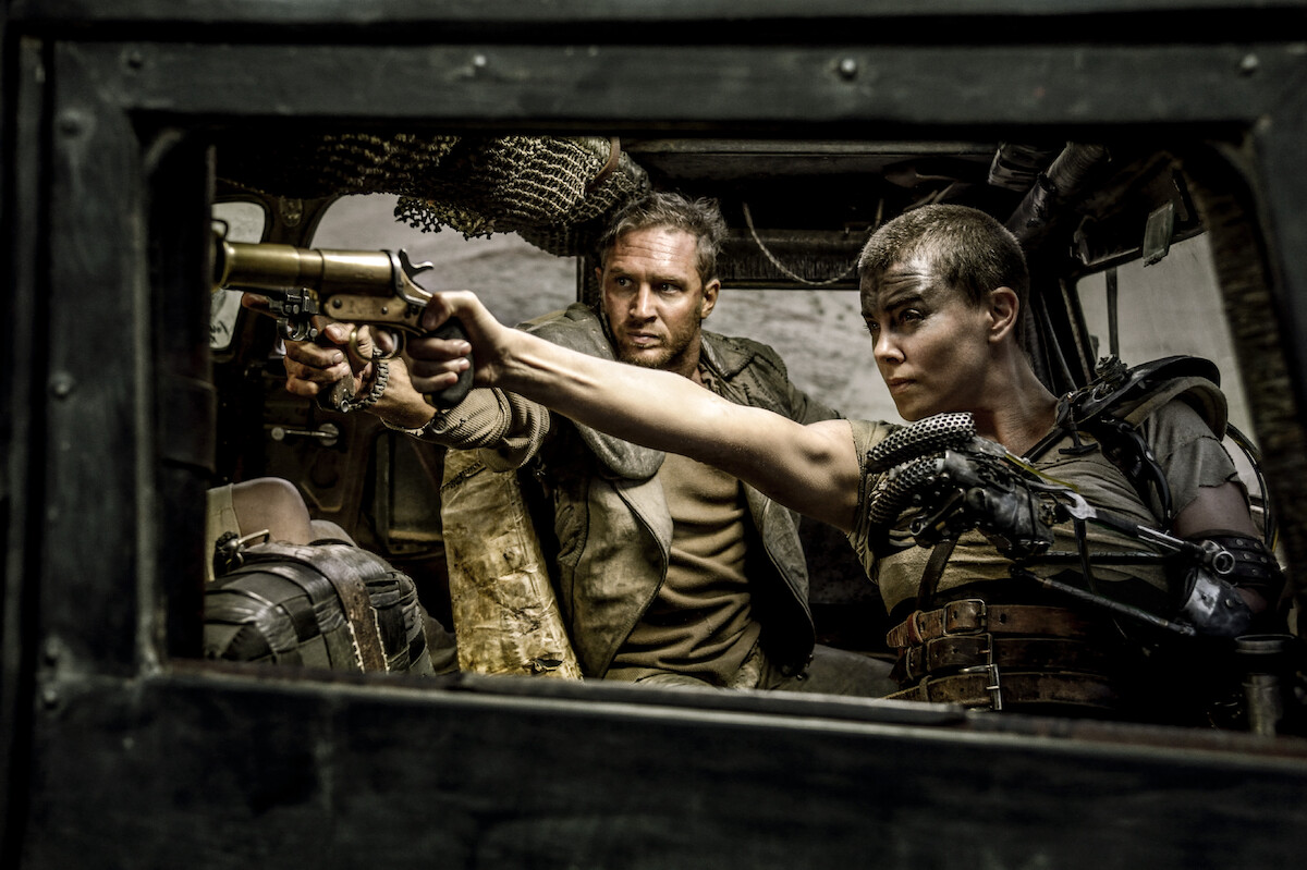 Charlize Theron as Imperator Furiosa and Tom Hardy as Max Rockatansky in ‘Mad Max: Fury Road’ driving in a truck while shooting out the passenger side window.