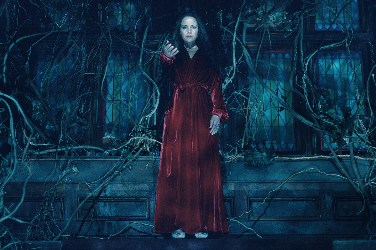 Woman in a long red dress stands in a dark, eerie room with stained glass windows and twisted, overgrown vines, reaching her hand forward as if summoning or warding off something.