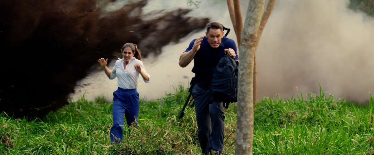 John Cena as Mason and Alison Brie as Claire run through smoke in a production still from ‘Freelance.’