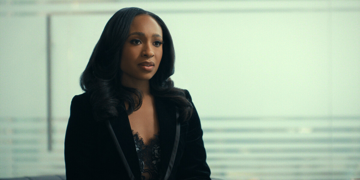 Woman with long dark hair in a black blazer sits indoors, with a blurred modern office background and horizontal lines visible through glass, appearing thoughtful or engaged in conversation.