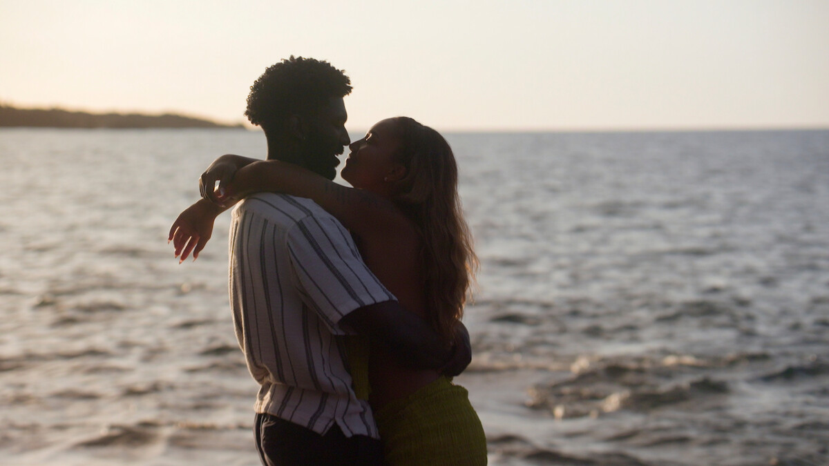 A couple embraces affectionately on a beach at sunset, standing close together with the ocean and a distant shoreline in the background.