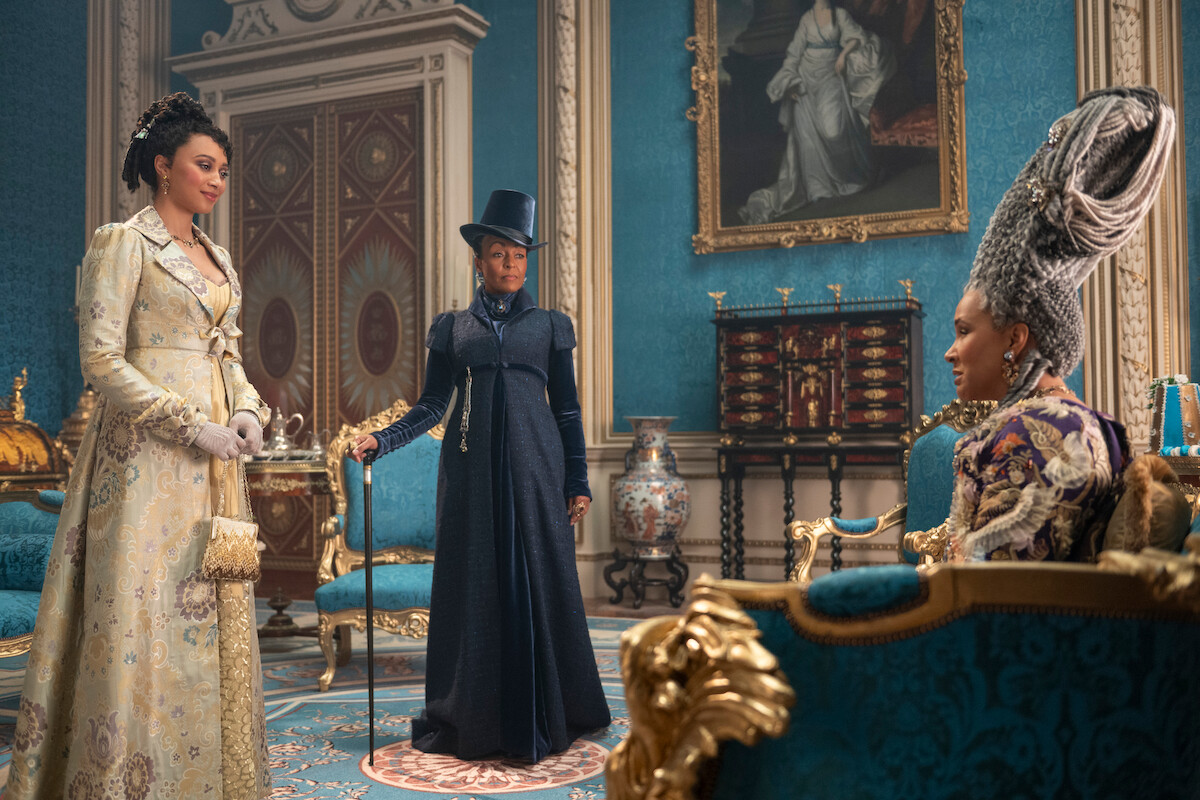 Três mulheres vestidas com trajes históricos sofisticados conversam em um suntuoso salão azul e dourado, com decoração real e pinturas clássicas, sugerindo um ambiente aristocrático ou palaciano.