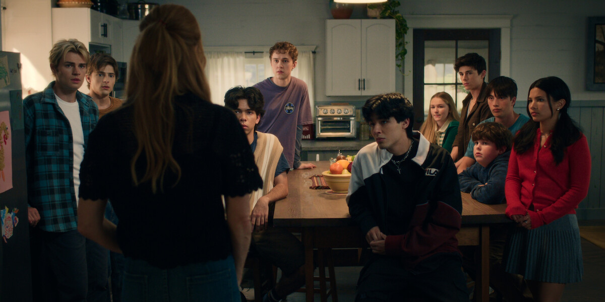 A woman stands facing a group of teenagers gathered around a kitchen table in a cozy, warmly lit home, all focused on her as if listening intently to an important conversation.