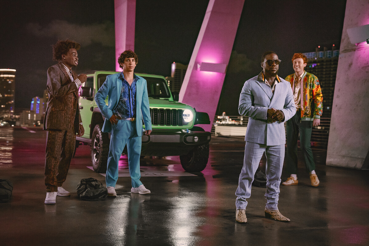 Four men in colorful suits stand on a rooftop at night beside a green Jeep, illuminated by pink and purple lights with a city skyline in the background.