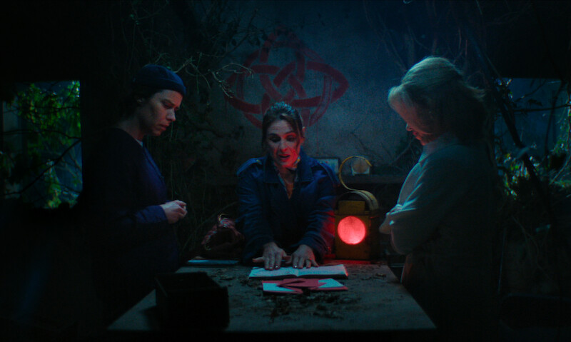 Three people gather around a dimly lit table with red symbols in a mysterious, overgrown room, discussing or examining papers, creating a tense and secretive mood. A strange diagram is drawn on the wall behind them.