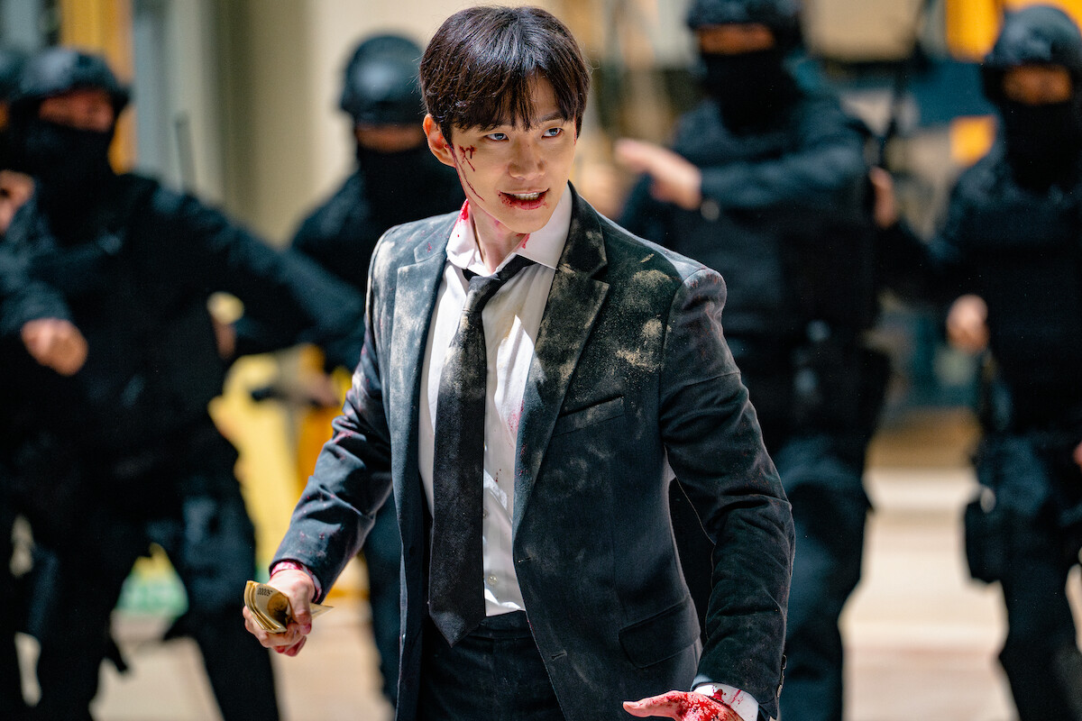 Man in a bloodied suit stands defiantly holding an object, face bruised, surrounded by armed SWAT officers in black uniforms. Scene appears tense and dramatic, suggesting action or conflict in an indoor environment.