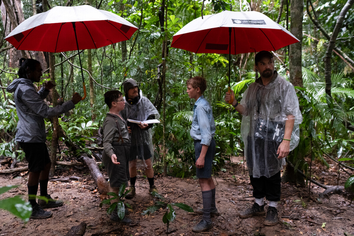 David McKenna as Piggy, Director Marc Munden, and Lox Pratt as Jack stand in the middle of a densely wooded tropical forest in the rain rehearsing a scene for Lord of the Flies under large red umbrellas.