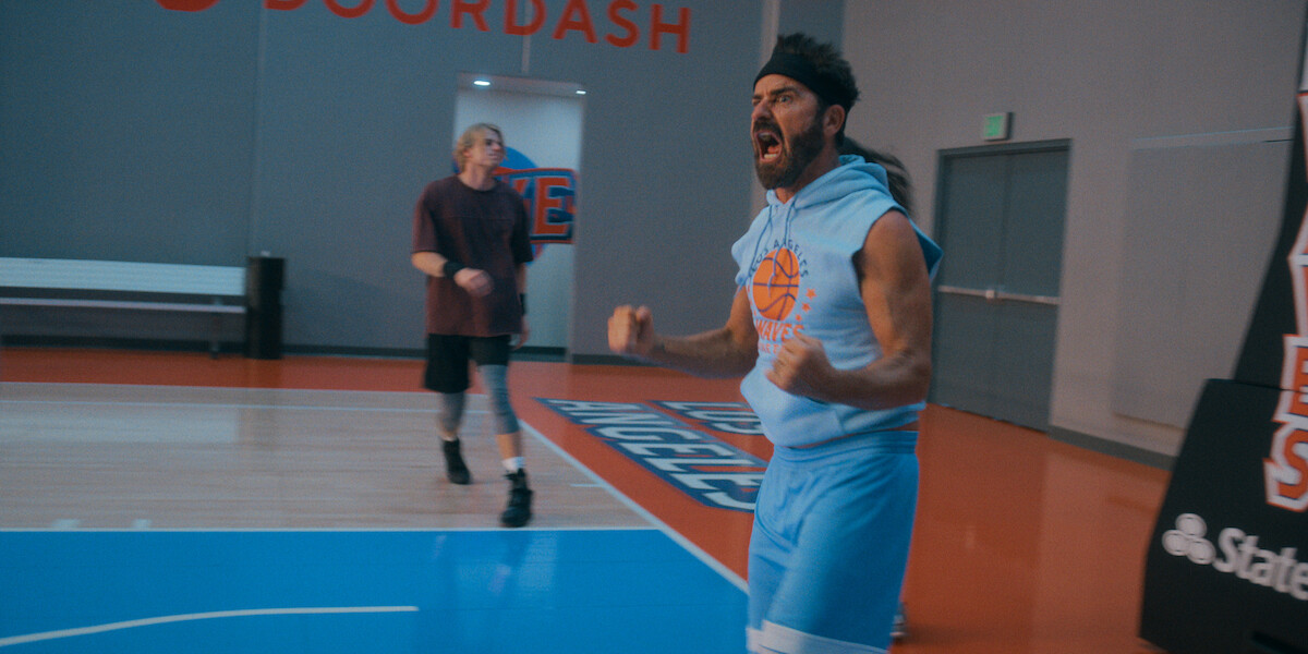Excited man in blue basketball outfit celebrates on an indoor court while another player walks behind him; basketball court features branding and seating in a gymnasium setting.