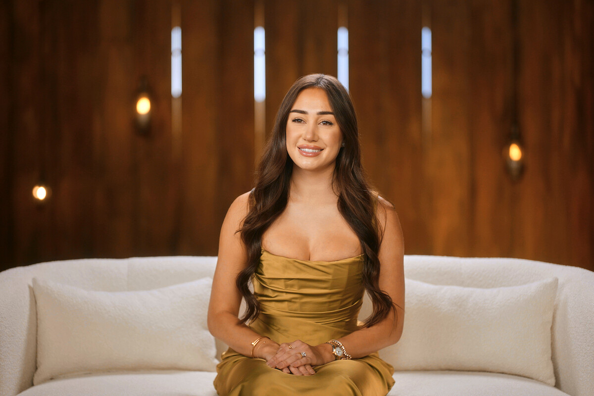 Woman with long brown hair in a gold dress sits on a white couch, smiling, against a warm wooden background with soft decorative lights and vertical slits, in a modern indoor setting.