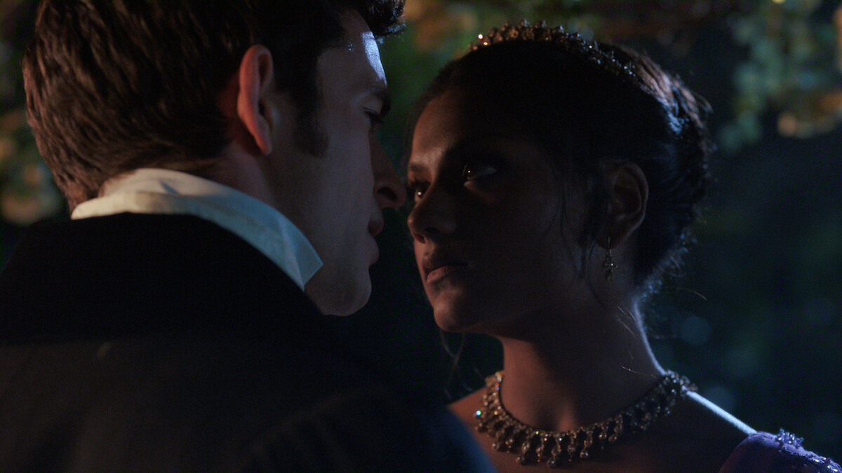 Two people dressed in formal, historical attire stand closely facing each other in a dimly lit outdoor setting at night, creating an intimate and dramatic atmosphere.
