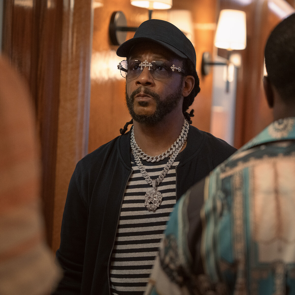 Man wearing sunglasses, cap, striped shirt, and thick chain necklace stands indoors in a warmly lit hallway, facing two other people whose backs are visible in the foreground.