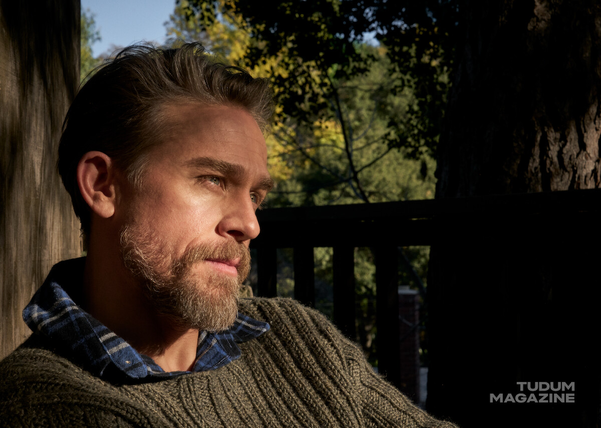 Charlie Hunnam sits on a porch wearing a green sweater and blue plaid shirt underneath, sunlight illuminating his blue eyes.