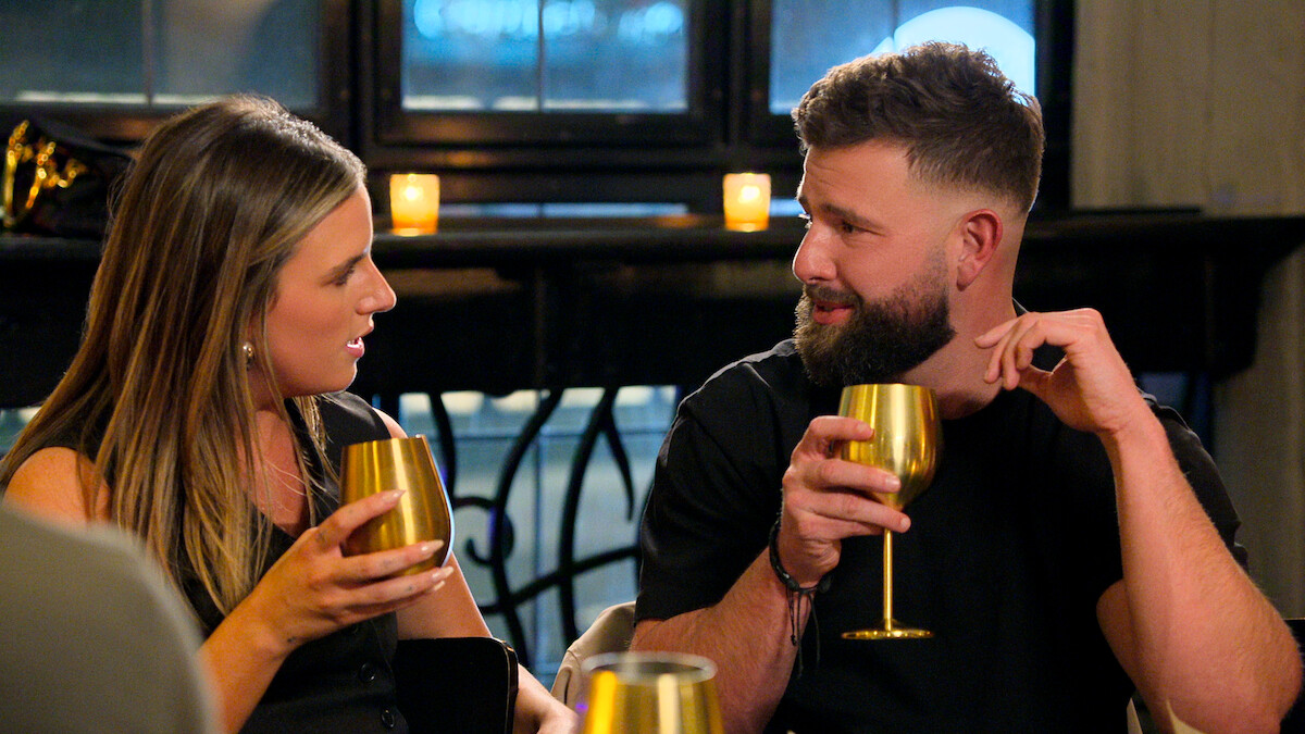 A woman and man sit indoors at a table holding gold wine glasses, having a conversation in a cozy, dimly lit setting with candles in the background.