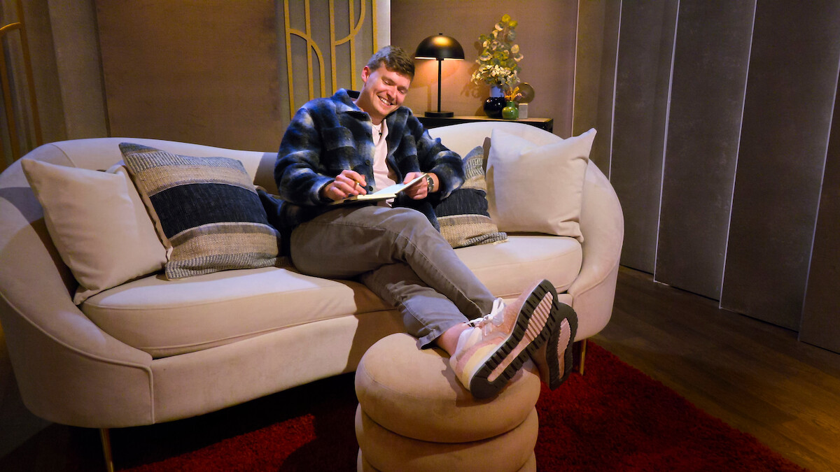 Smiling person lounging on a cream sofa with pillows, writing in a notebook, feet up on an ottoman, in a cozy living room with warm lighting, a lamp, and decorative flowers in the background.