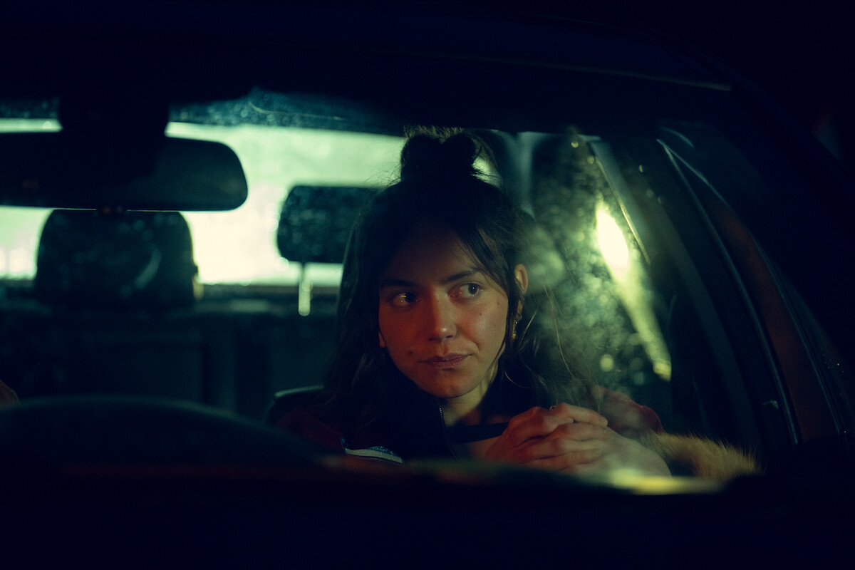 A woman sits alone in a dimly lit car at night, illuminated by moody lighting with rain on the window, giving a contemplative and introspective atmosphere.