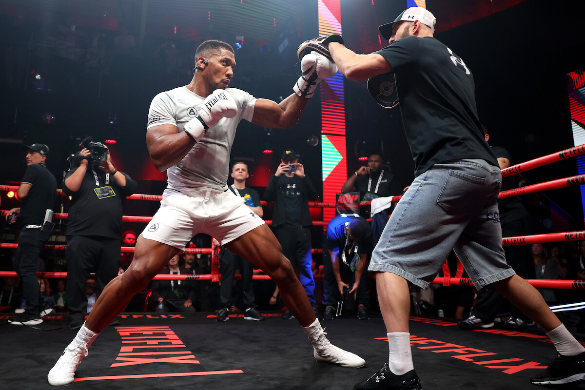 Anthony Joshua in white sports gear trains in a boxing ring, punching mitts held by a coach in casual clothes, with a crowd of spectators and photographers watching in a dimly lit, energetic gym environment.