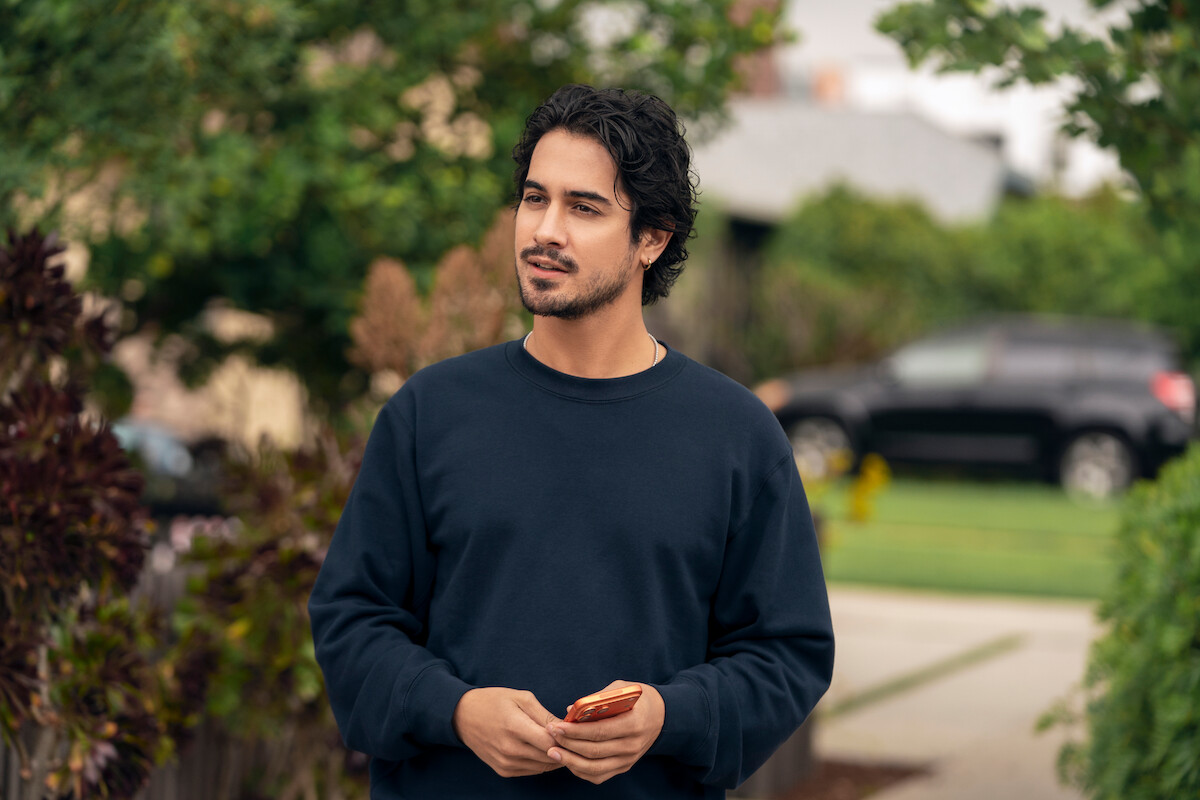 Man in a dark sweater holding a phone stands outdoors in a suburban neighborhood with trees, bushes, and a parked car in the background.