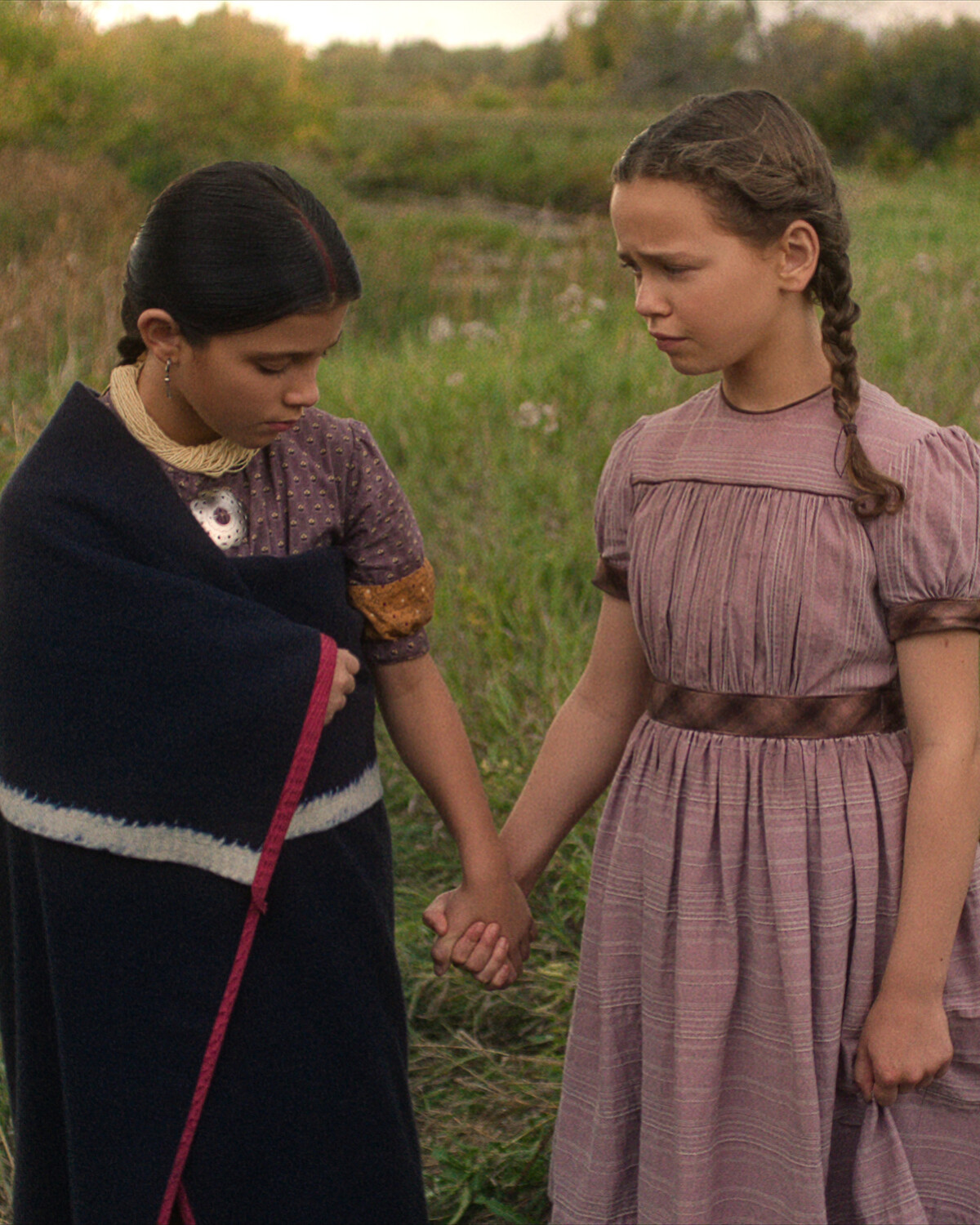 Two young girls in vintage dresses stand in a grassy field holding hands; one wears a dark shawl and looks down, while the other, with braided hair, looks at her with concern. The setting appears rural and peaceful.