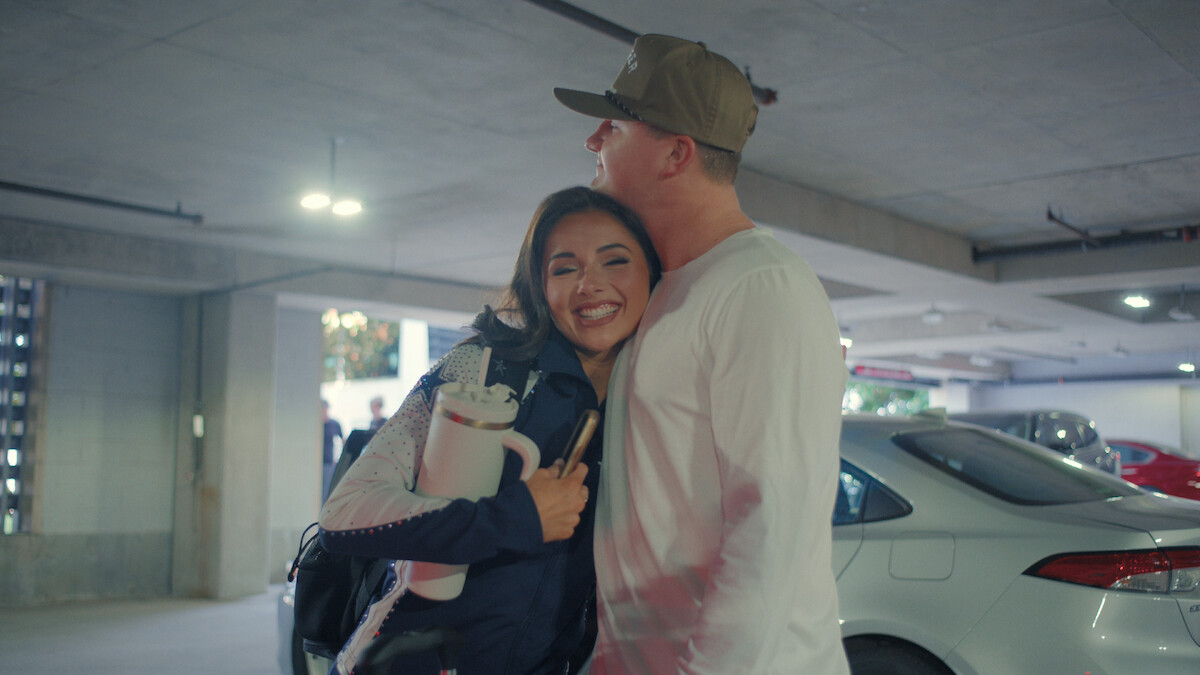 Two people hug and smile in a parking garage, surrounded by parked cars under artificial lighting, suggesting a warm reunion or farewell in an urban setting.