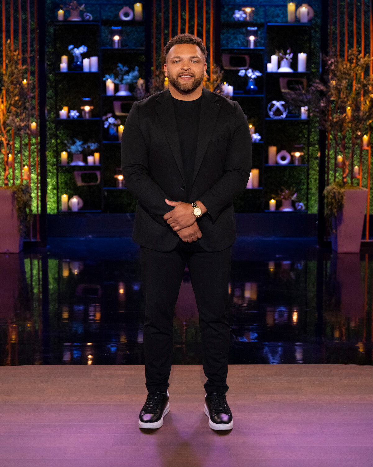 A man in black suit and sneakers stands on a studio stage with shelves filled with candles, plants, and decorative items behind him, warm lighting, and a stylish, modern setting.